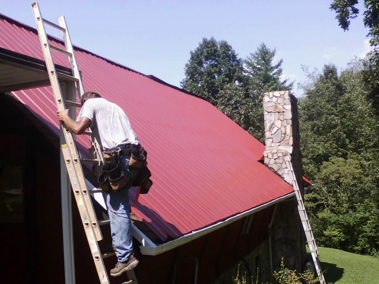 Roofer on a ladder beside a red metal roof and stone chimney — Soffit & Fascia Repair by Roofing Brothers in Amador City
